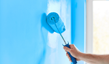 Hand using a paint roller to apply bright blue paint to a wall near a window.