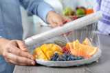 Putting plastic food wrap over plate of fresh fruits and berries at wooden table