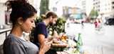 woman sitting at a café looking at her smartphone. Man in the backround is eating. out of focus, bicycles are passing by on the street.
