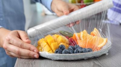 Putting plastic food wrap over plate of fresh fruits and berries at wooden table