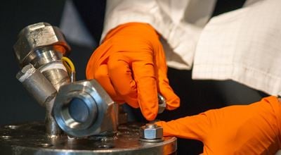 Person wearing orange gloves working with a metal bolt on industrial equipment.